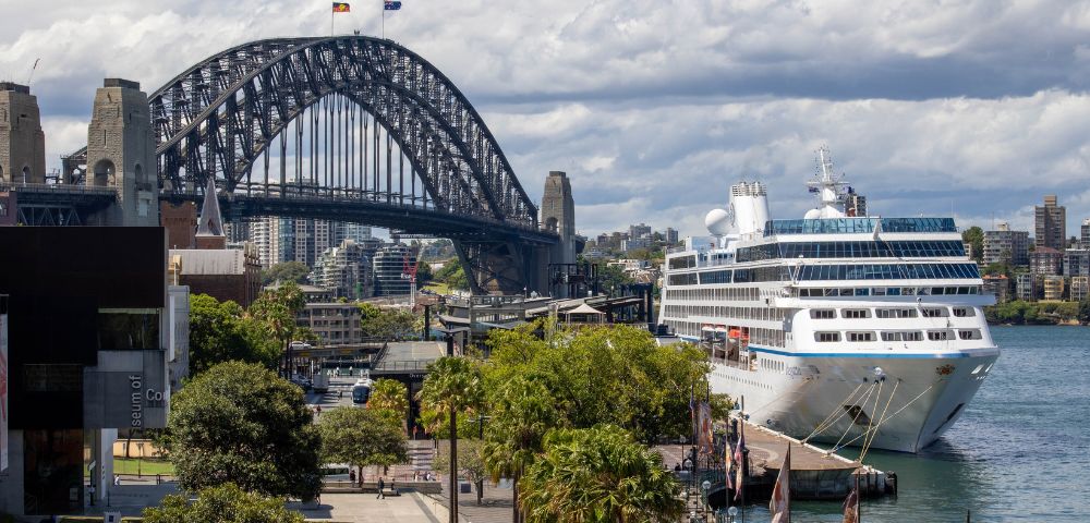A large white cruise ship is docked in a harbor with the Sydney Harbour Bridge in the background and the city skyline beyond.