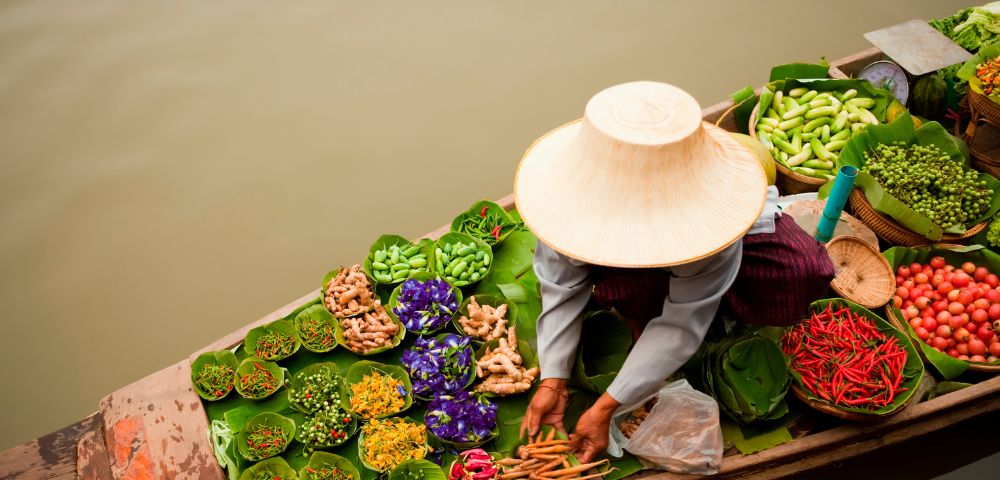 A vendor in a conical hat arranges fresh produce, including red chilies, ginger, purple flowers, and green vegetables, in a boat on a body of water.