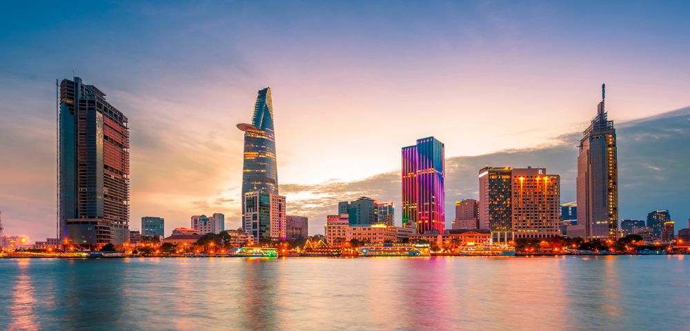 Panoramic view of a modern city skyline at dusk, with numerous skyscrapers of varying heights and architectural styles illuminated by warm lights, reflected in the calm waters of a river in the foreground.
