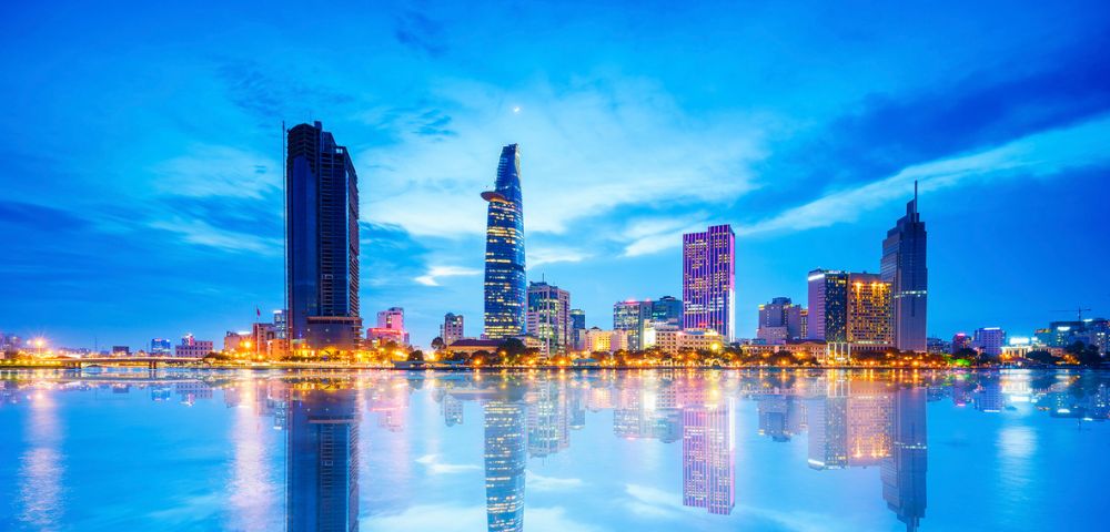 Panoramic view of a modern city skyline at twilight, with illuminated skyscrapers reflecting in a calm body of water.