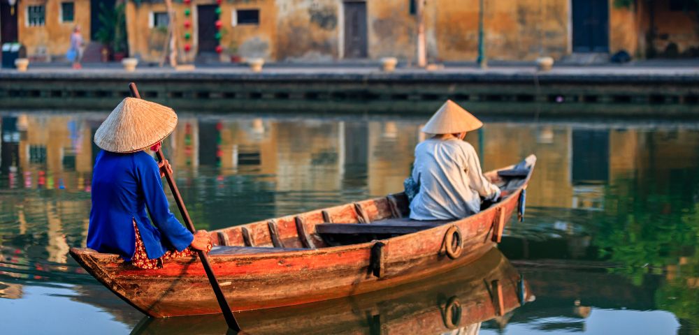 Two individuals wearing conical hats navigate a wooden boat down a calm river, with buildings reflected in the water.