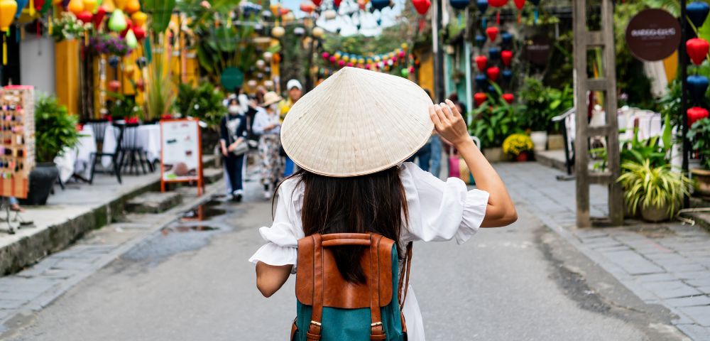 A person wearing a conical hat and a white ruffled shirt walks down a cobblestone street lined with shops and greenery, with other pedestrians in the blurred background.
