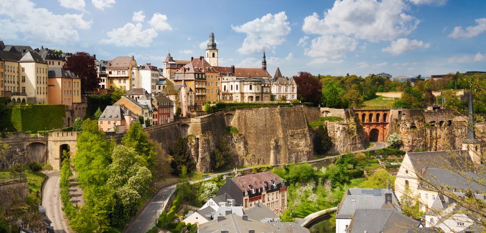 Panoramic view of Luxembourg City's historic buildings atop cliffs with lush greenery and blue sky with clouds.