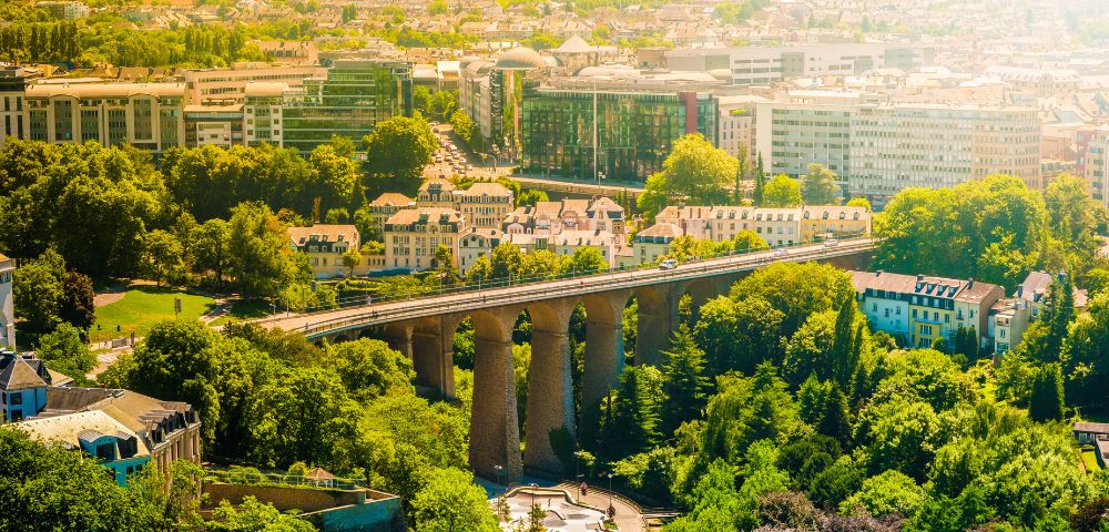 Aerial view of a city with a stone arch bridge crossing over lush green trees and residential buildings in bright sunlight.