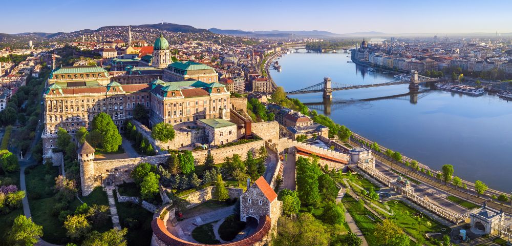 Aerial view of Budapest, showcasing the historic Buda Castle with green domes, the Chain Bridge, and the Danube River under a clear blue sky.