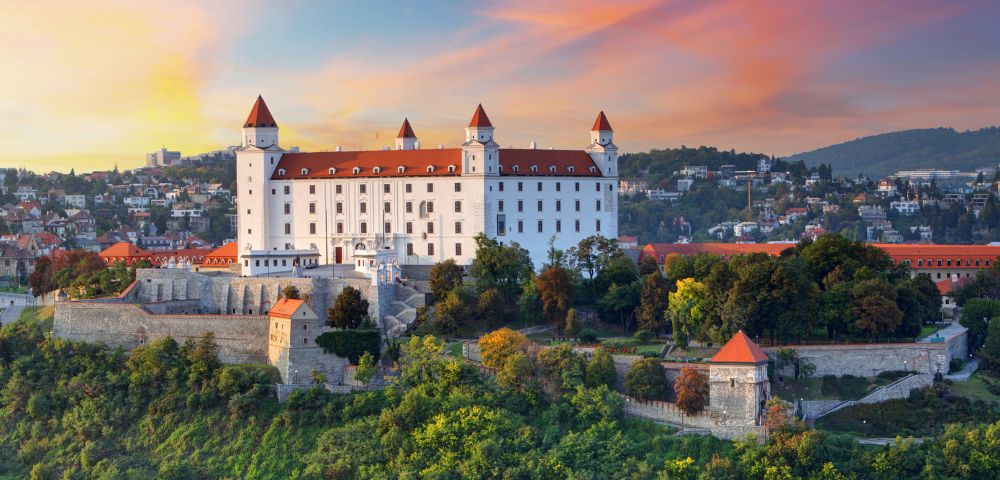 Bratislava Castle in Slovakia at sunset, with a warm sky and lush greenery. The white castle has red rooftops, surrounded by trees and city buildings.