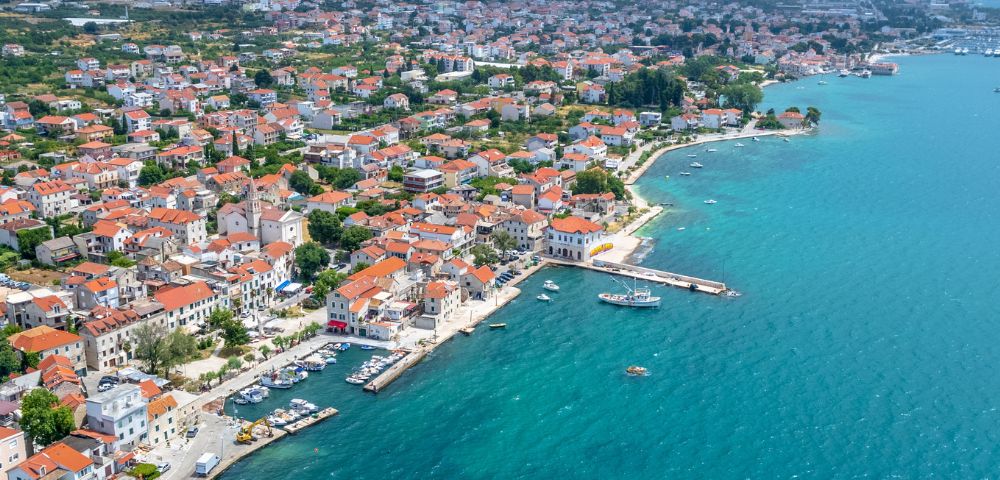 Aerial view of a coastal town with red-roofed buildings, small boats docked at piers, and clear blue sea along the shoreline.