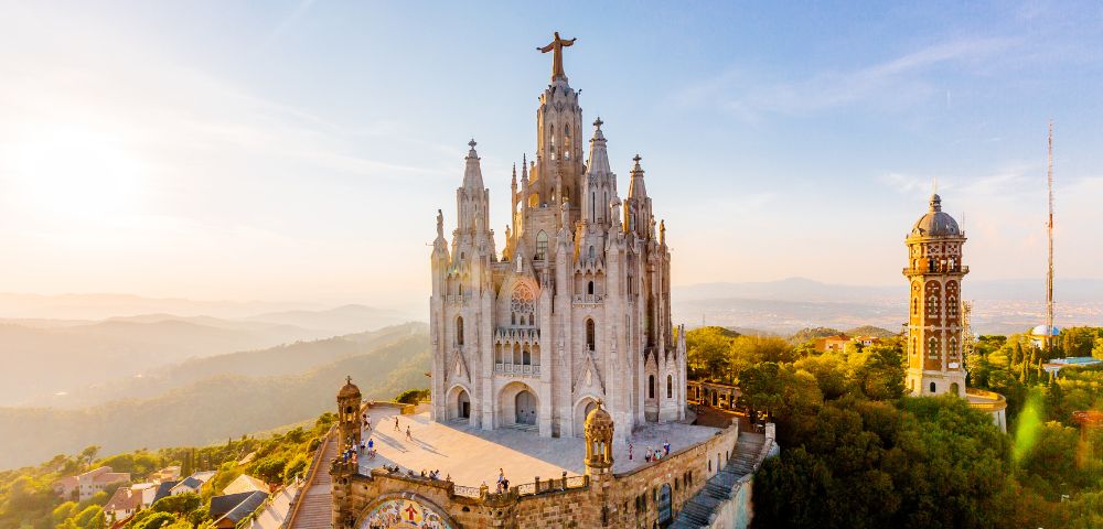 A majestic hilltop basilica with a large Jesus statue on top, overlooking a scenic landscape. The sky is clear and sunny, conveying peace and grandeur.
