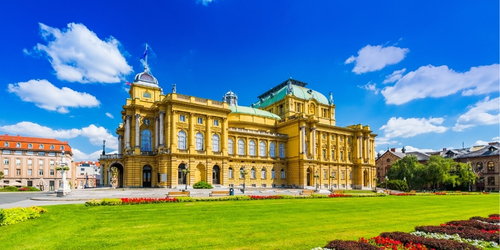 A grand, historic building with ornate architecture under a clear blue sky, surrounded by vibrant green lawns and colorful flowerbeds, conveys elegance and tranquility.