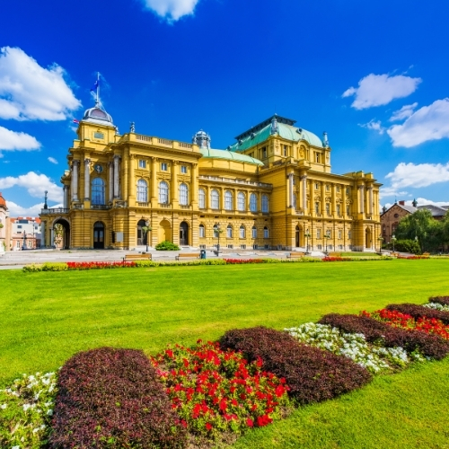A grand, historic building with ornate architecture under a clear blue sky, surrounded by vibrant green lawns and colorful flowerbeds, conveys elegance and tranquility.