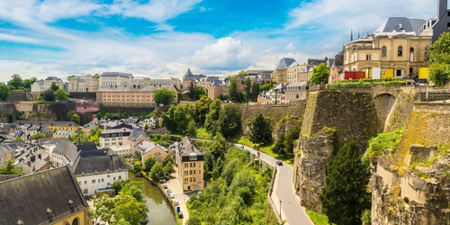 Scenic view of Luxembourg City with historic buildings, lush greenery, a river, and a bright blue sky with clouds.