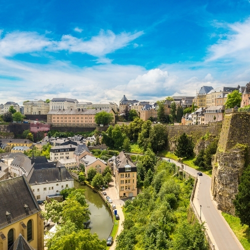 Scenic view of Luxembourg City with historic buildings, lush greenery, a river, and a bright blue sky with clouds.