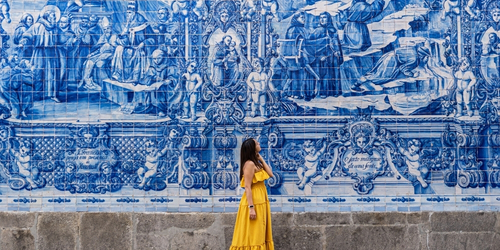 Woman in a yellow dress stands before a large blue and white ornate tile mural depicting religious scenes.