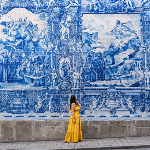 Woman in a yellow dress stands before a large blue and white ornate tile mural depicting religious scenes.