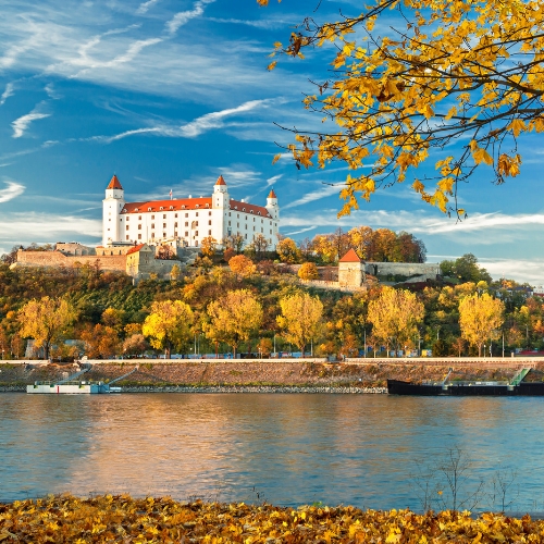 Majestic white castle with red roofs atop a hill, surrounded by vibrant autumn trees. A calm river flows beneath a bright blue sky with wispy clouds.