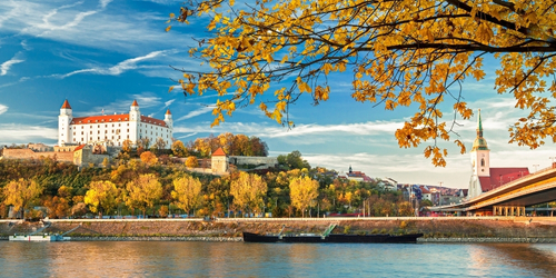 Majestic white castle with red roofs atop a hill, surrounded by vibrant autumn trees. A calm river flows beneath a bright blue sky with wispy clouds.