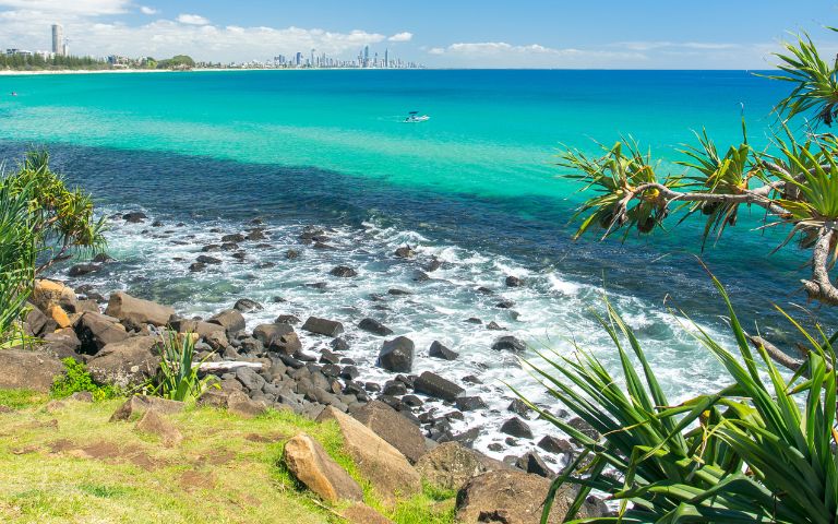 A bright, sunny coastal scene with turquoise and deep blue ocean water. Rocky shorelines in the foreground transition to a sandy beach with a distant city skyline under a clear blue sky. Tropical foliage frames the right side of the image.