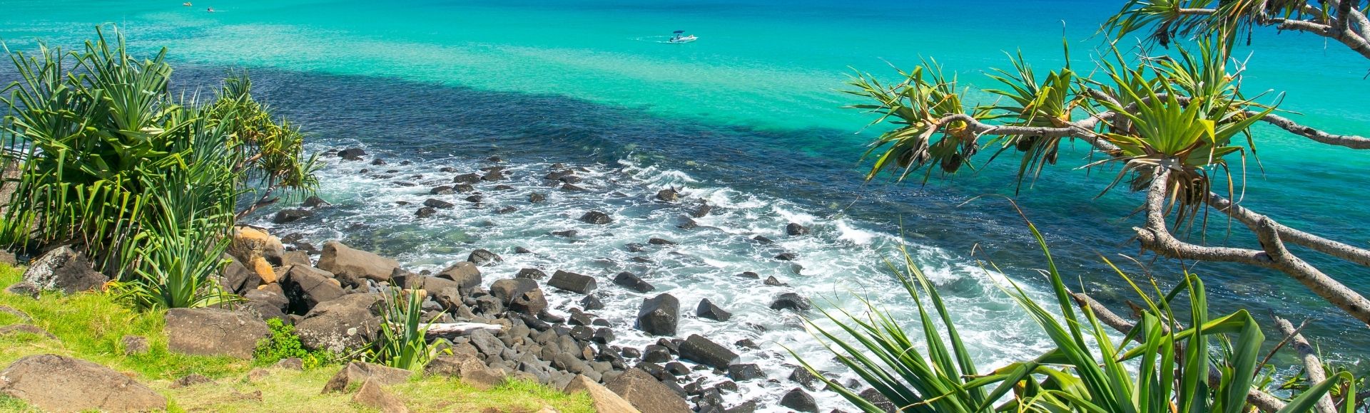 A bright, sunny coastal scene with turquoise and deep blue ocean water. Rocky shorelines in the foreground transition to a sandy beach with a distant city skyline under a clear blue sky. Tropical foliage frames the right side of the image.