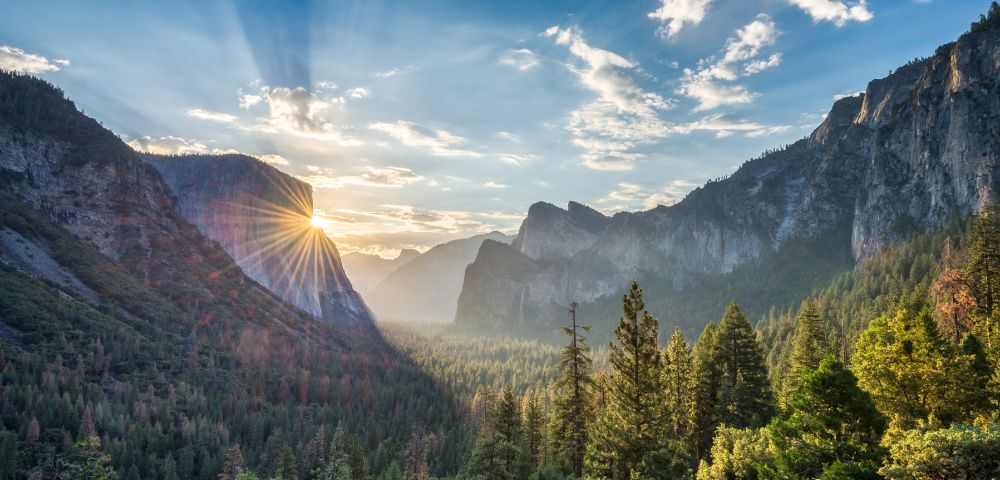 A sunburst shines over the granite cliffs of Yosemite Valley, illuminating a dense evergreen forest below under a bright blue sky with scattered clouds.