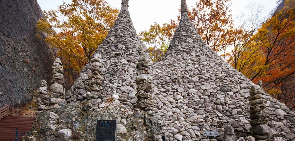 Two conical stone structures situated among colorful autumn trees. The structures are densely covered with rocks, with a plaque visible in front of them. A pathway leading up to the site can be seen in the left background.