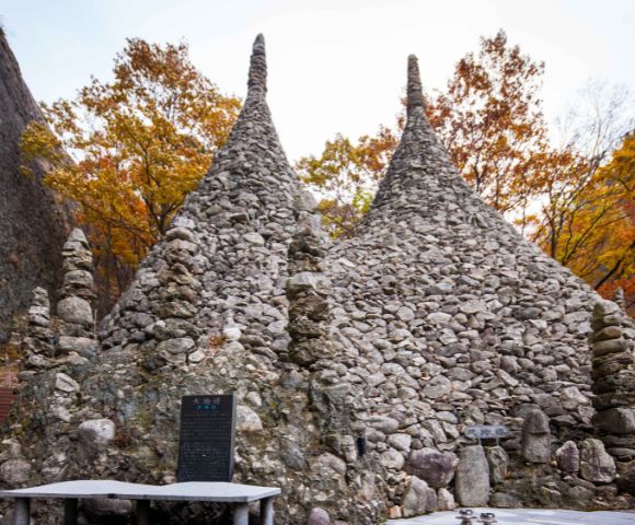 Two conical stone structures situated among colorful autumn trees. The structures are densely covered with rocks, with a plaque visible in front of them. A pathway leading up to the site can be seen in the left background.