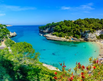 A picturesque turquoise bay with sailboats, a sandy beach lined with sunbathers, and lush green cliffs under a clear blue sky.