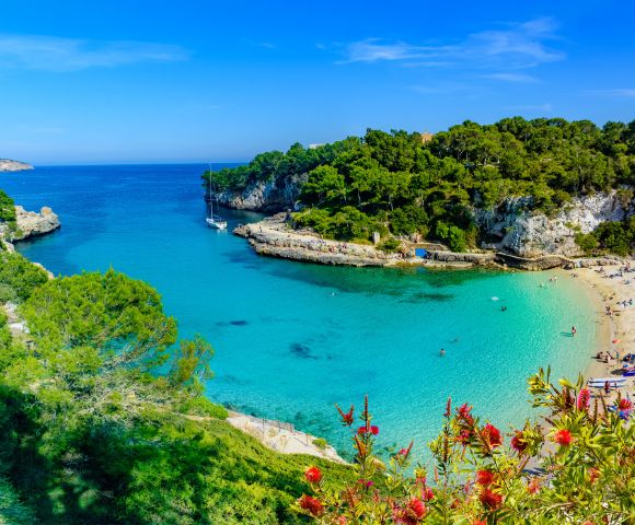 A picturesque turquoise bay with sailboats, a sandy beach lined with sunbathers, and lush green cliffs under a clear blue sky.