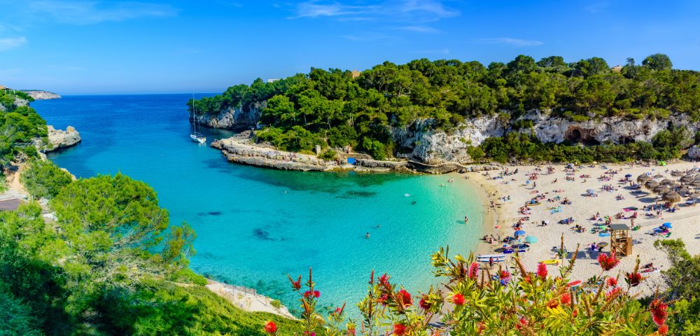 A picturesque turquoise bay with sailboats, a sandy beach lined with sunbathers, and lush green cliffs under a clear blue sky.
