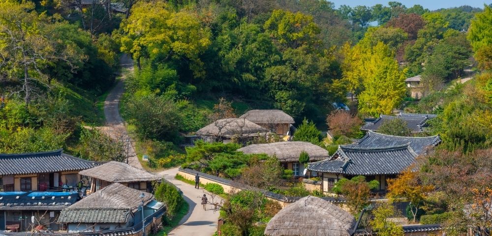 Aerial view of a traditional village featuring several thatched-roof houses along winding paths. The landscape is green with trees and ornamental shrubs, creating a serene environment typical of historic architecture.