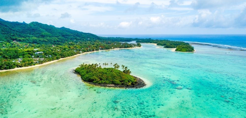 Aerial view of a small, green island surrounded by clear turquoise waters near a larger tropical coastline under a partly cloudy sky.