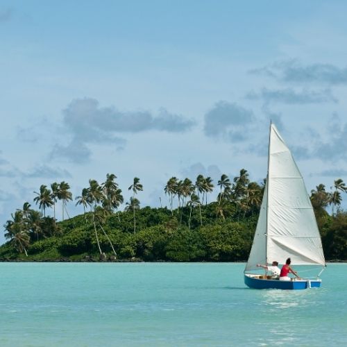 Sailboat with two people glides on turquoise water near a lush island with tall palm trees under a partly cloudy sky.