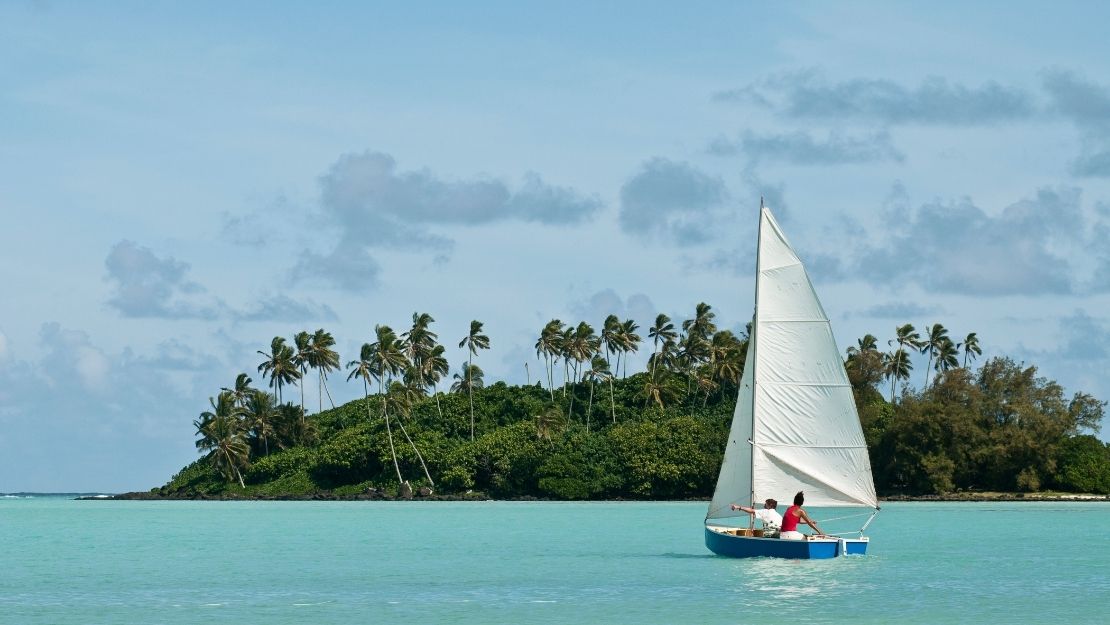 Sailboat with two people glides on turquoise water near a lush island with tall palm trees under a partly cloudy sky.