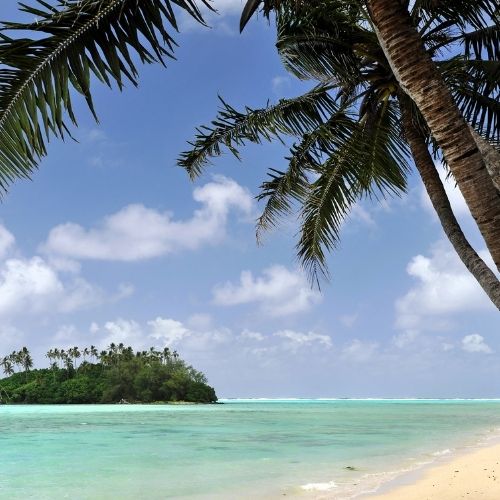 A tropical island with palm trees in the foreground and background, set against a bright blue sky with scattered clouds. The clear, turquoise water gently laps at a sandy beach.