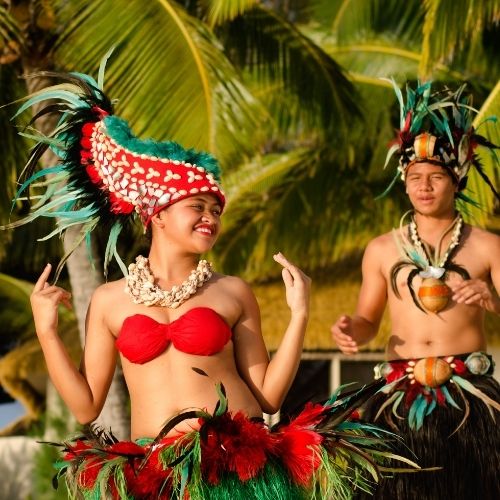 A woman and a man in traditional Polynesian attire, performing a dance against a tropical backdrop.