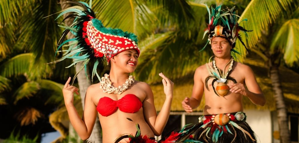 A woman and a man in traditional Polynesian attire, performing a dance against a tropical backdrop.