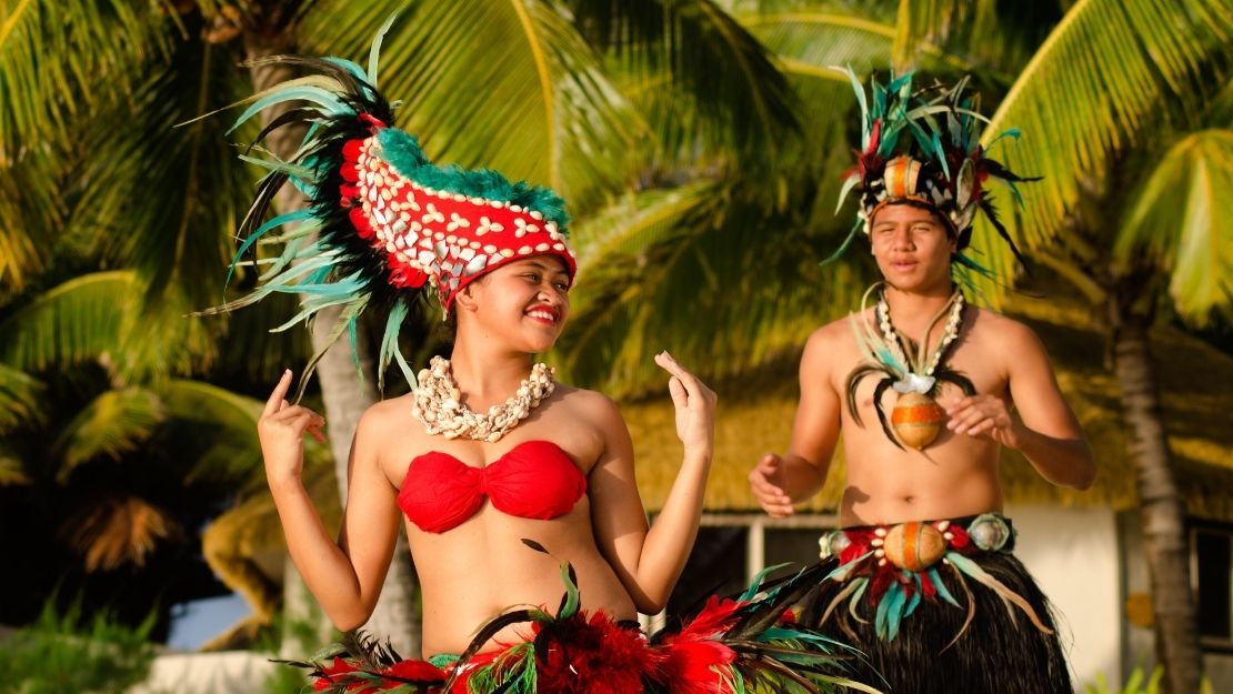 A woman and a man in traditional Polynesian attire, performing a dance against a tropical backdrop.
