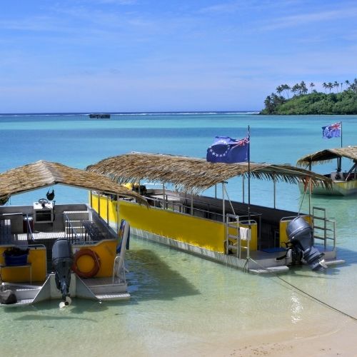 Boats with thatched roofs anchored in a calm bay with clear blue water and palm trees in the background.