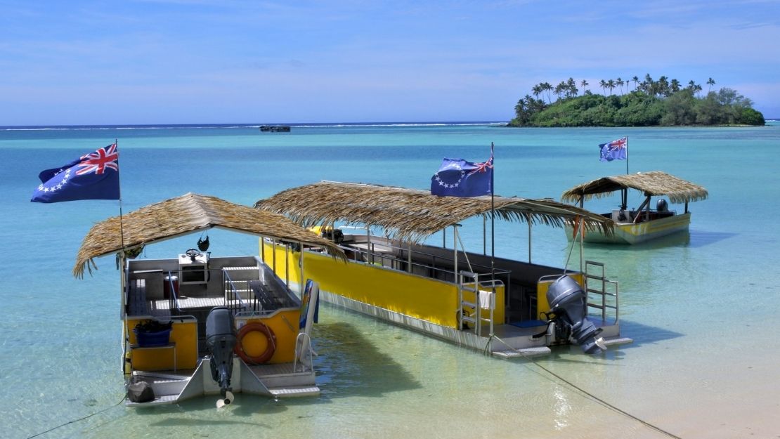 Boats with thatched roofs anchored in a calm bay with clear blue water and palm trees in the background.