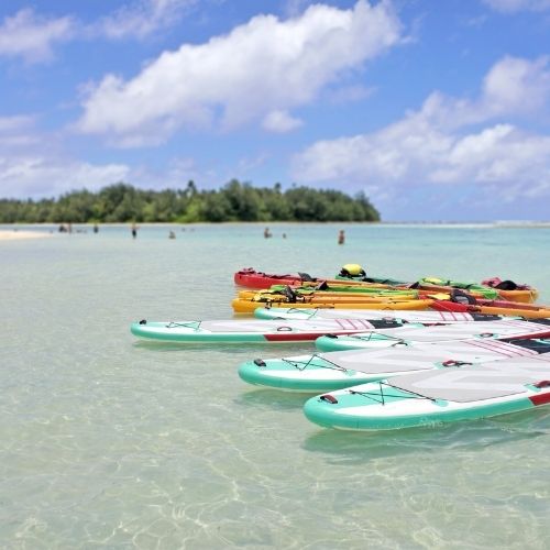 Kayaks and paddleboards lined up on a clear, calm beach with a tropical island in the background.