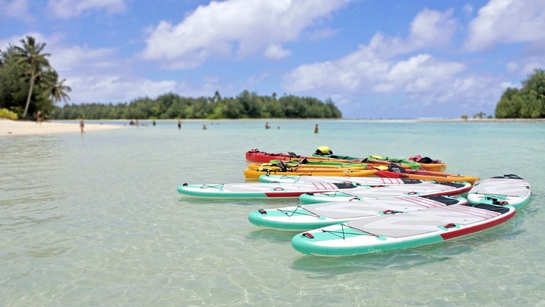 Kayaks and paddleboards lined up on a clear, calm beach with a tropical island in the background.