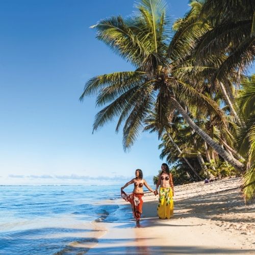 Two women walking hand in hand along a scenic beach, surrounded by palm trees and clear blue water.