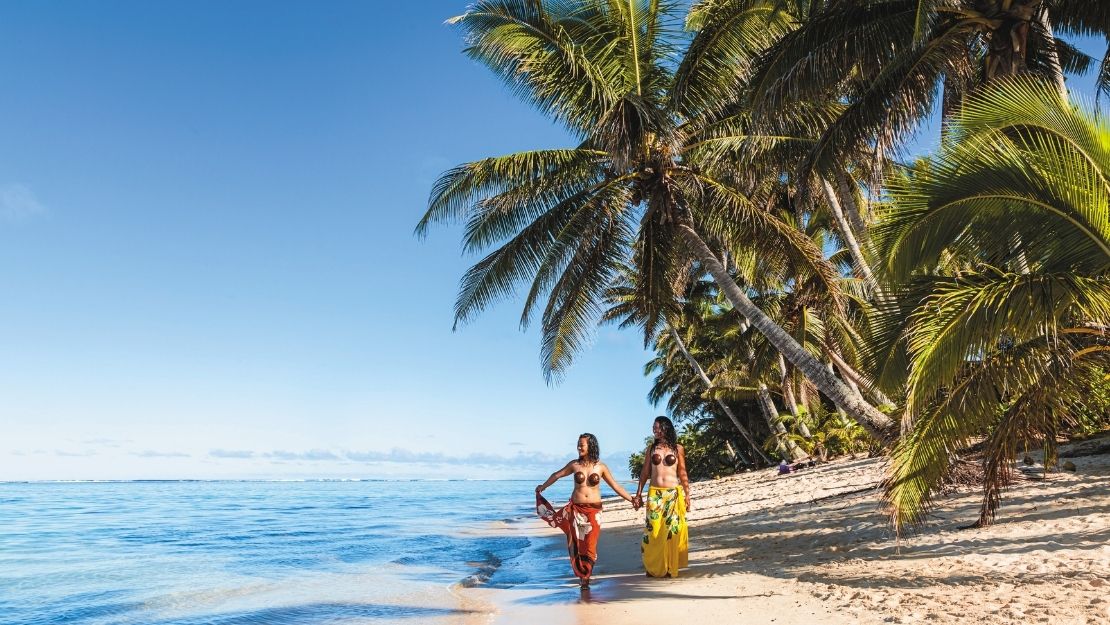 Two women walking hand in hand along a scenic beach, surrounded by palm trees and clear blue water.