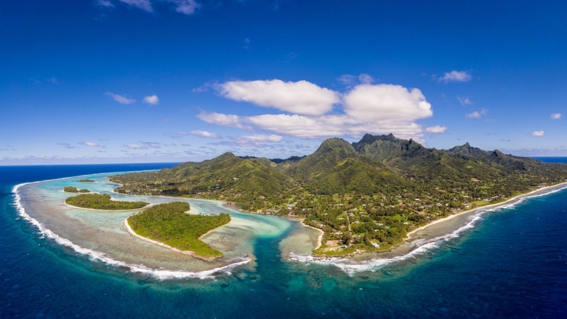 Aerial view of a tropical island with a sandy beach, turquoise waters, and a sailboat anchored nearby, surrounded by palm trees under a blue sky with scattered clouds.