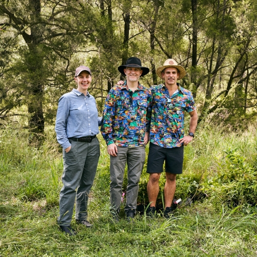 Three people smiling and standing outdoors in a grassy, wooded area, two wearing colorful shirts and hats, one in a blue shirt and cap.