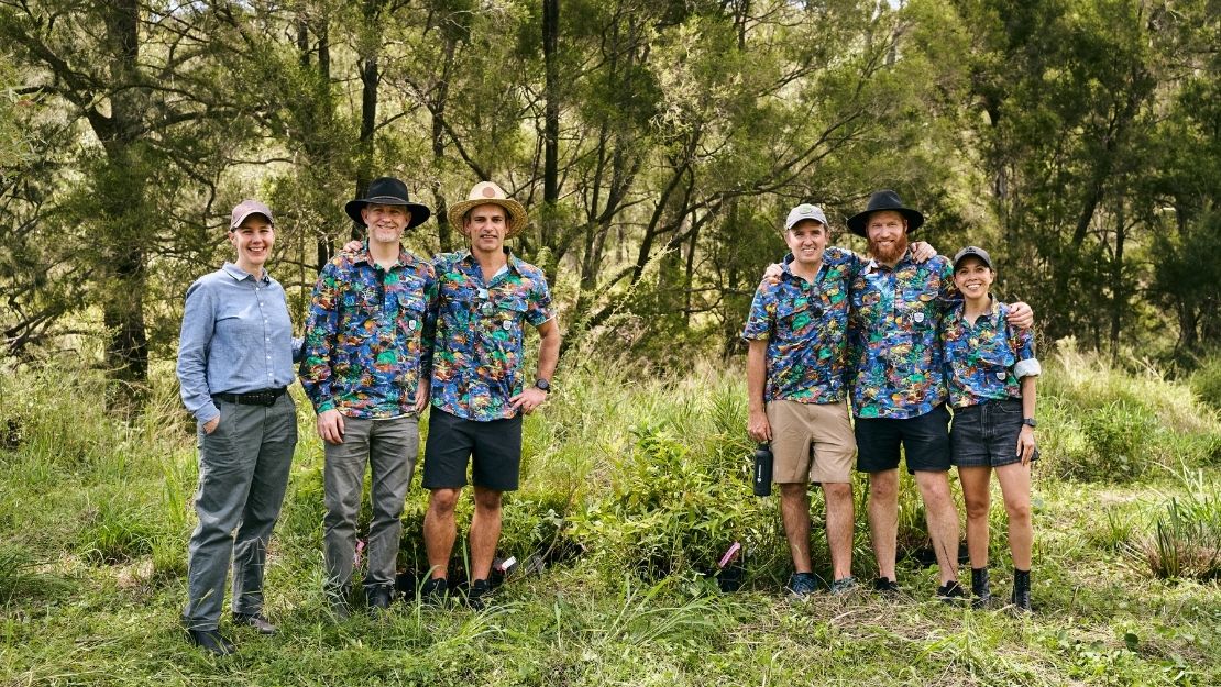 Six people standing outdoors on grass in front of trees, five wearing colorful patterned shirts, two wearing hats, smiling at the camera.