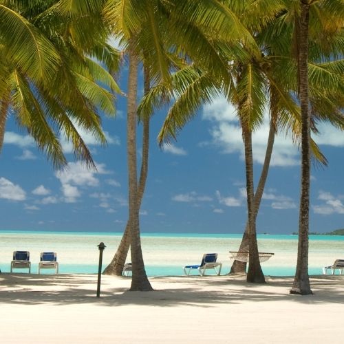 A tropical beach scene with tall palm trees framing a white sand beach, turquoise water, and a clear blue sky with scattered clouds. Lounge chairs are visible on the sand near the water's edge.