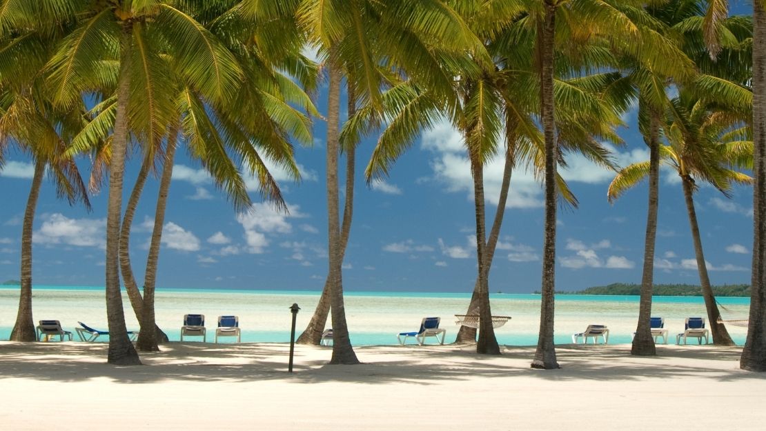 A tropical beach scene with tall palm trees framing a white sand beach, turquoise water, and a clear blue sky with scattered clouds. Lounge chairs are visible on the sand near the water's edge.