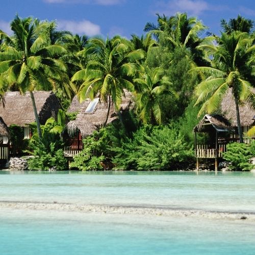 Tropical huts with thatched roofs nestled among palm trees along a clear, shallow turquoise lagoon under a blue sky.