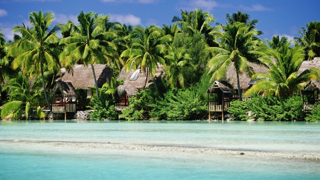 Tropical huts with thatched roofs nestled among palm trees along a clear, shallow turquoise lagoon under a blue sky.