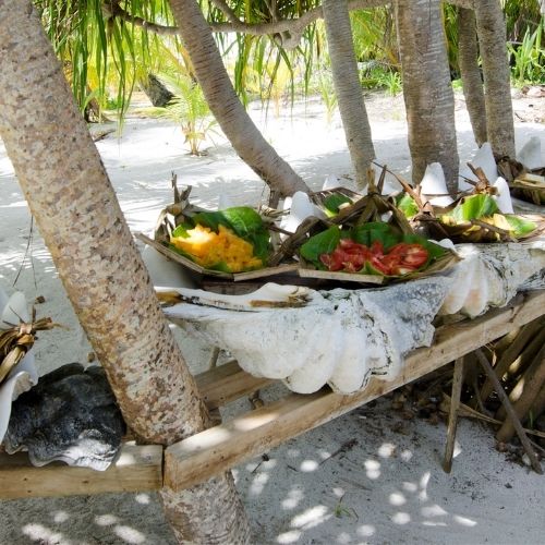 Tropical outdoor setting with wooden table holding plates of fresh fruit and large seashells under palm trees on sandy ground.